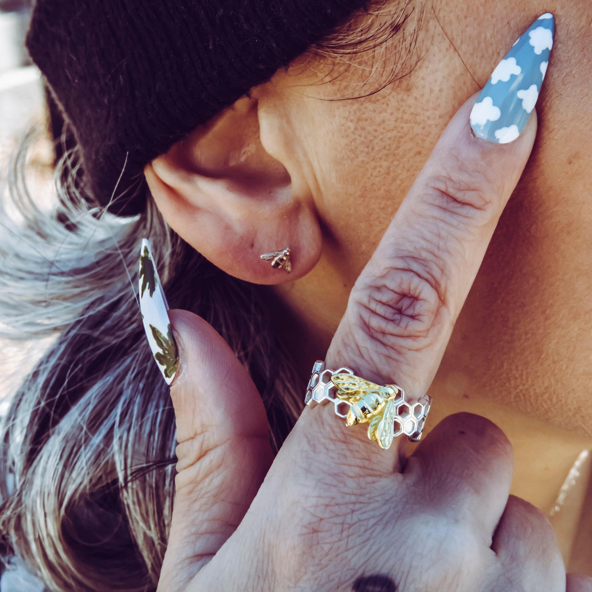 person with index finger to temple showcasing bee honeycomb ring. their ear is visible showing off 14K gold baby bee stud