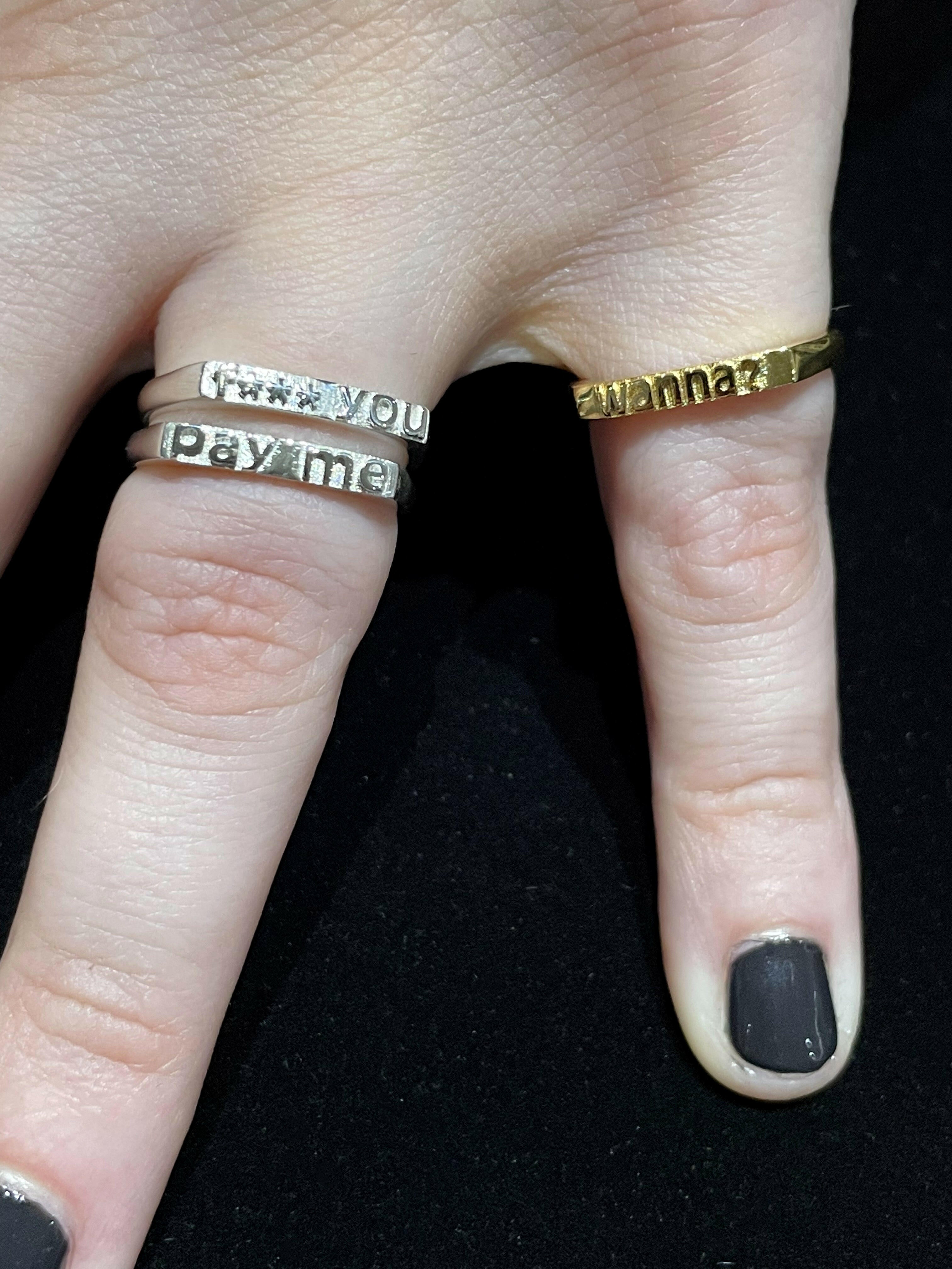 Two fingers, ring finger wearing 2 rings - first (top) 14 Karat white gold ring with text that reads "f*** you", second (bottom) 14 k white gold ring with text that reads "pay me". Pinky finger wearing a 14 karat yellow gold ring with text that reads "wanna?"