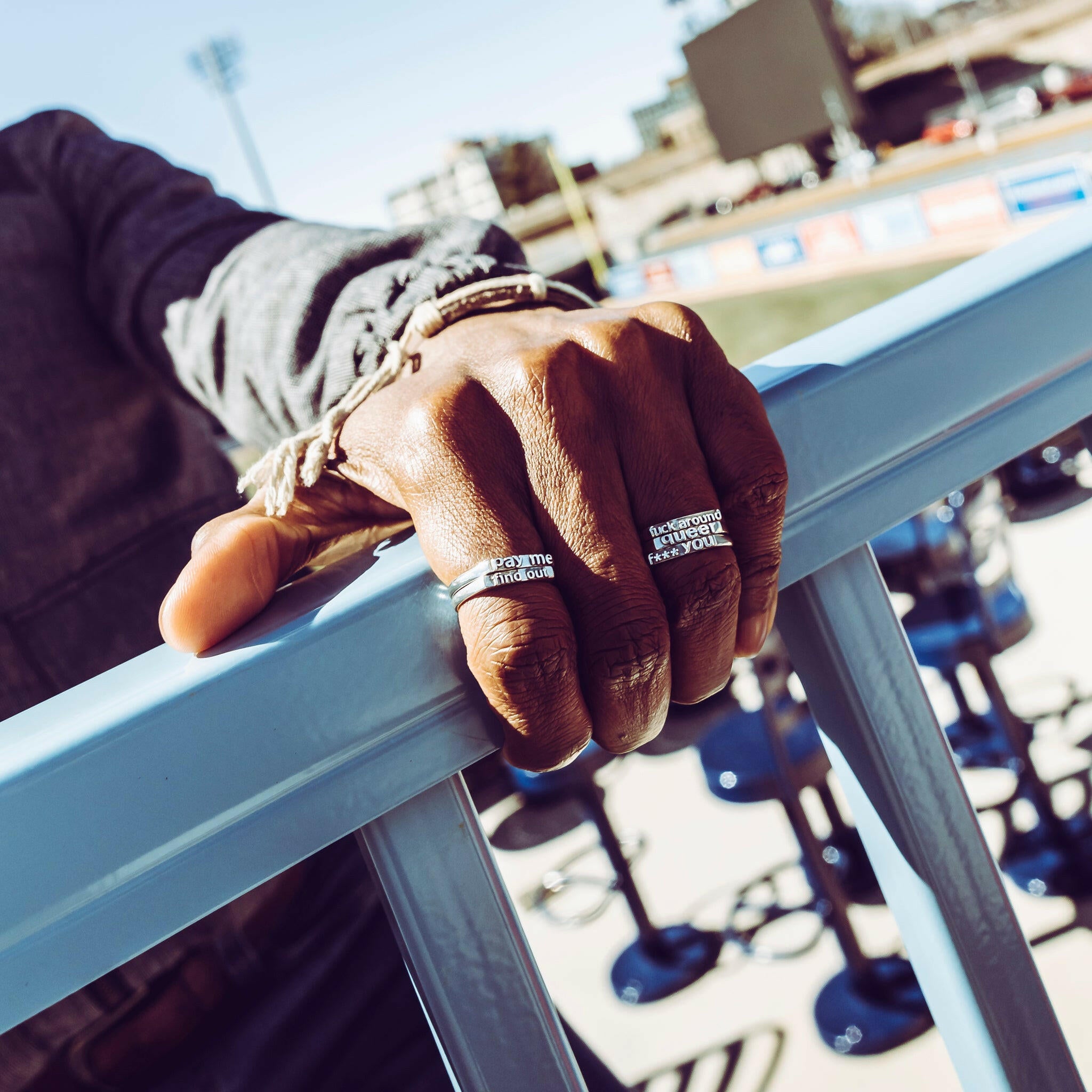 Hand holding a Railing displaying five Sterling Silver Word rings. Far Left (Index Finger)- two rings stacked atop one another, the top one being a ring that reads the text "Pay Me" the bottom ring reads "Find Out". Far Right (Ring Finger) Has 3 rings stacked on one another. The top rings reads text saying "Fuck Around". The middle ring has text that reads "queer". The bottom ring has text that reads "F*** you" a censored version of "fuck you"